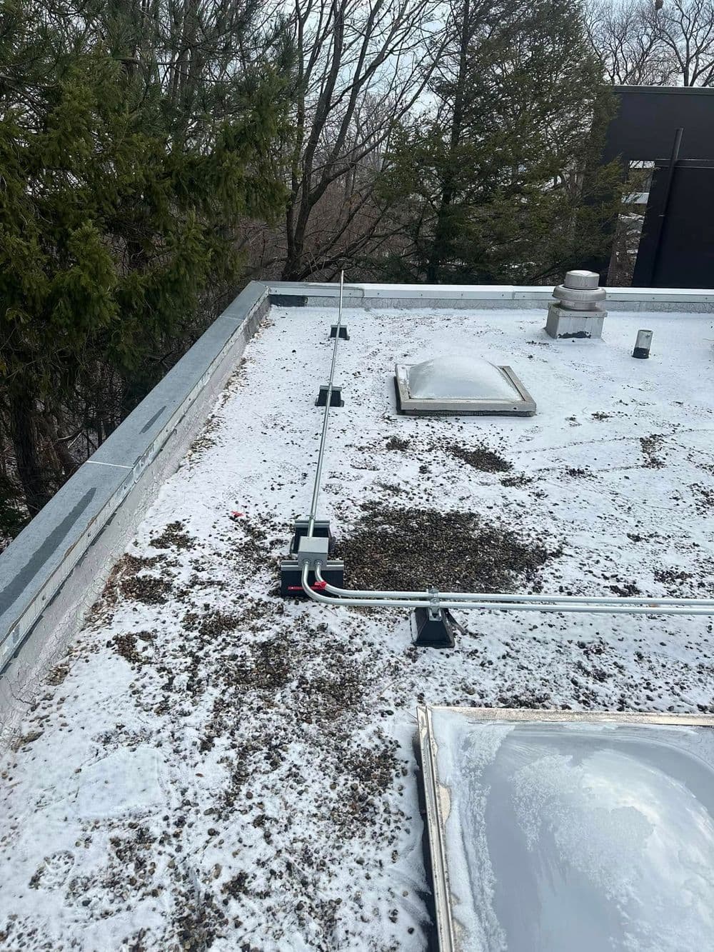 Roof top covered in snow with drainage pipes and a skylight visible. Trees in the background.