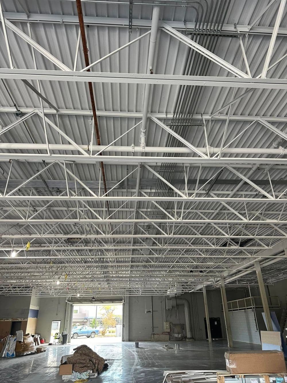 Interior of large commercial space showing a high ceiling with white trusses and exposed pipes.