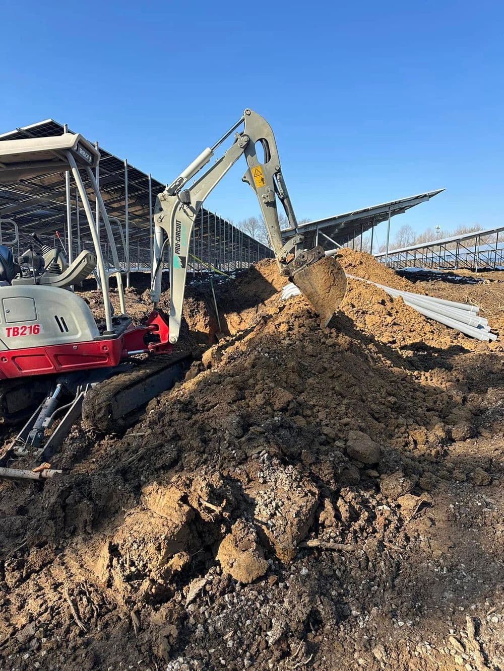 Excavator digging into a dirt mound at a construction site under a clear blue sky.
